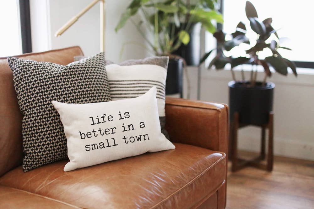 Life is Better in a Small Town Linen Pillow propped on a tan leather sofa alongside patterned black and white throw pillows, potted plants in the background