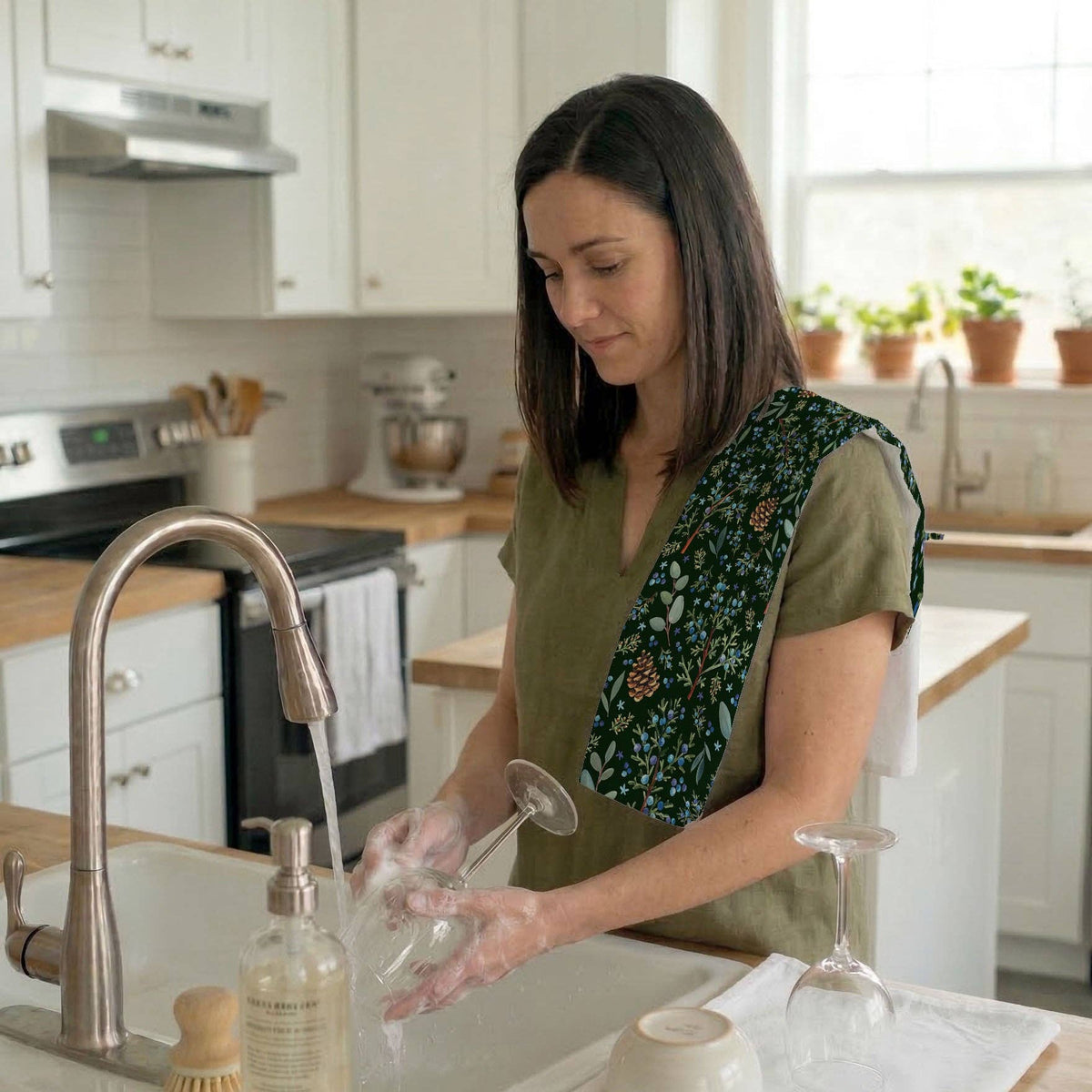 Winter Juniper Tea Towel tucked into an apron pocket while a person washes a wine glass at a kitchen sink