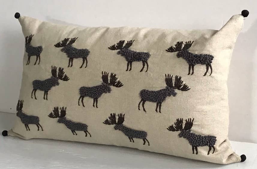Angled view of a beige cotton pillow with embroidered gray french-knot moose and black pom-poms at the corners, on white