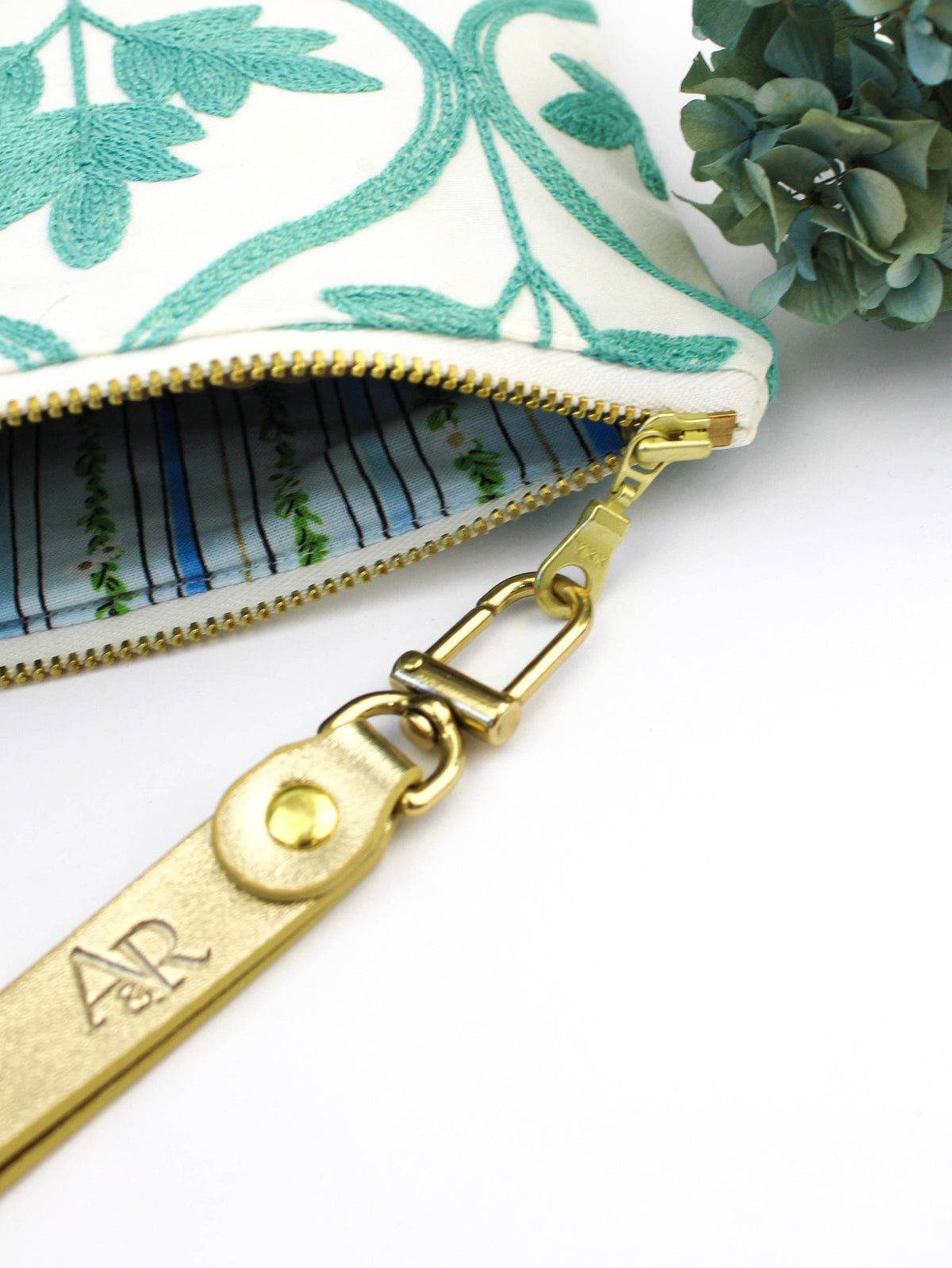Close-up of an open Mayfair Aqua Botanical wristlet purse showing a blue striped lining and gold zipper hardware, teal embroidered exterior