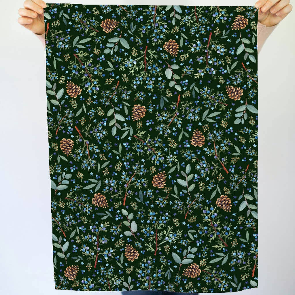 Winter Juniper Tea Towel held up fully by two hands showing the full pattern of juniper branches, blue berries, eucalyptus leaves, and golden pinecones