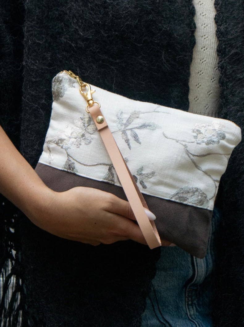 A woman holding a Winona Embroidered Linen wristlet, ivory linen with grey and metallic floral embroidery over a brown suede base, pink leather strap
