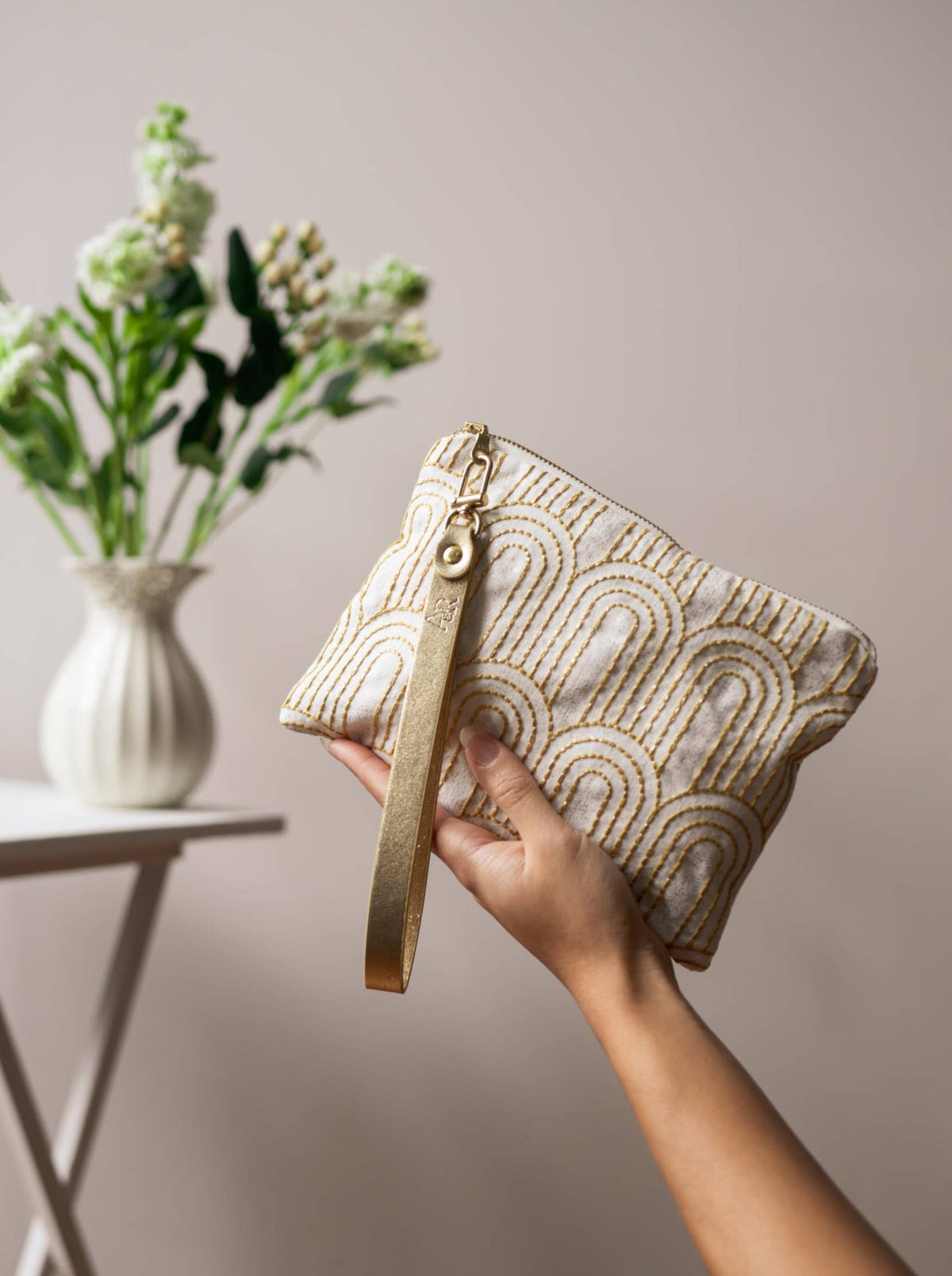A woman holding a Josephine Clutch wristlet purse by its gold strap, cream fabric with gold Art Deco arch embroidery, near a vase of white flowers