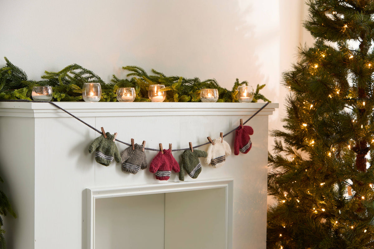 Mantel garland of tiny knit sweaters and hats in red, green, gray and cream beside a lit Christmas tree