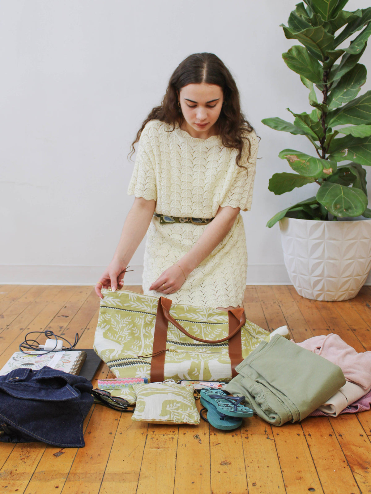 A woman packing a Folk Life Carry On Travel Bag and matching mini pouch with clothes, sandals, and a notebook, green and cream forest-motif jacquard fabric