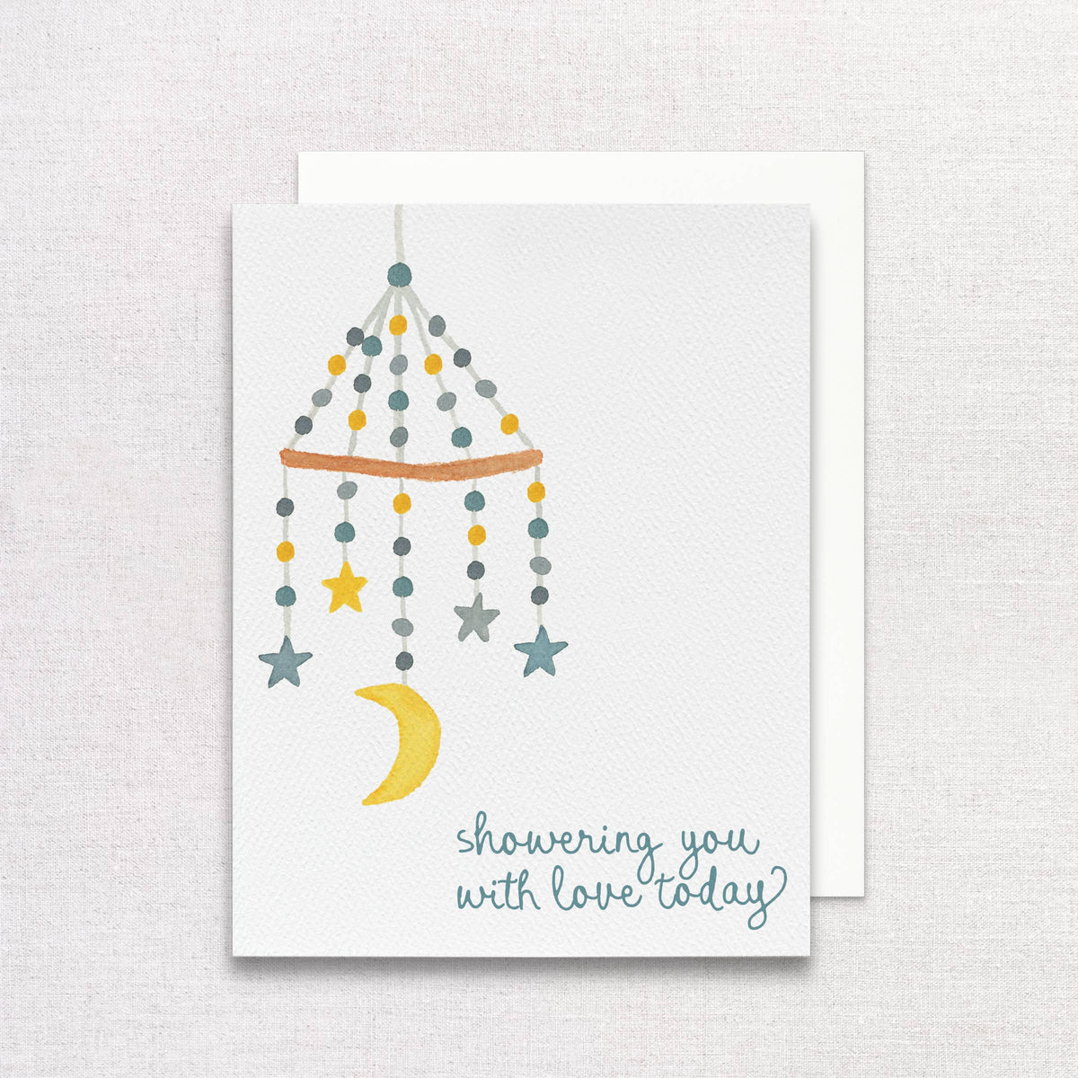 Showering You With Love card with a watercolor baby mobile of stars and a moon, on a white envelope over linen