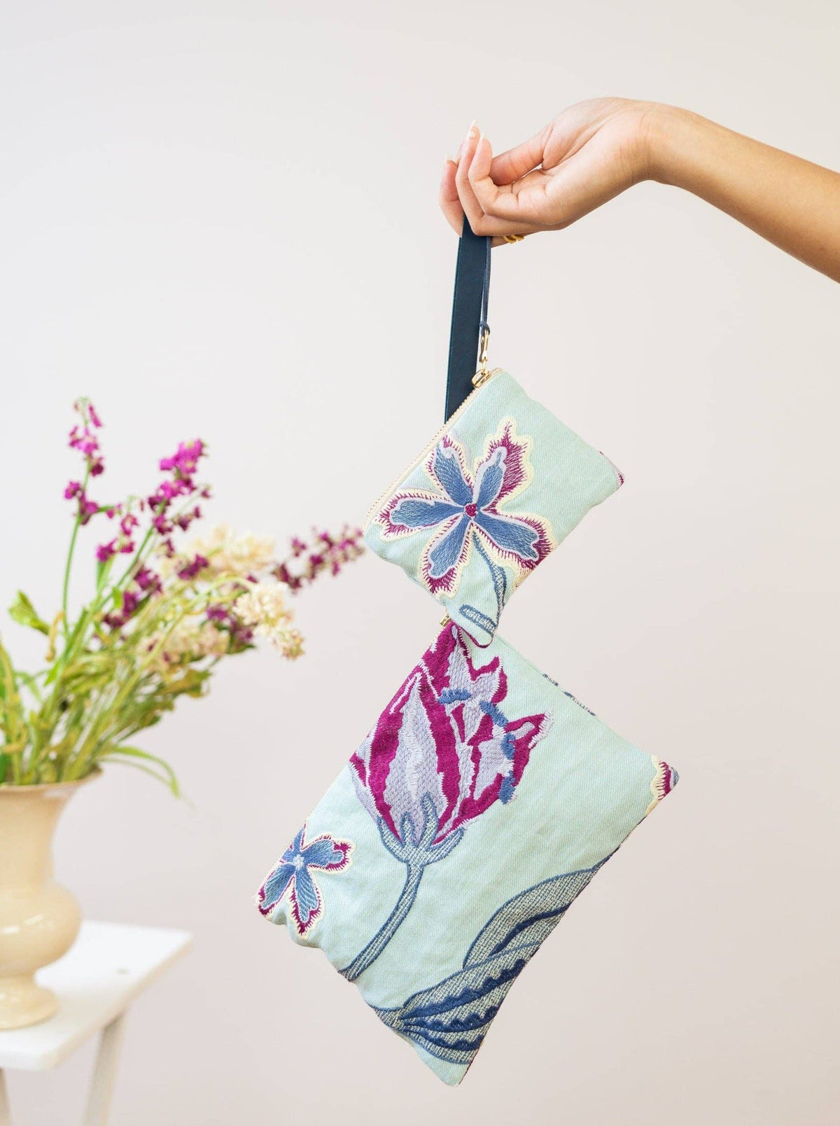 A woman holding an Enchanting Azure embroidered mini pouch and matching larger wristlet purse together by a navy strap, mint green fabric with magenta and blue floral embroidery