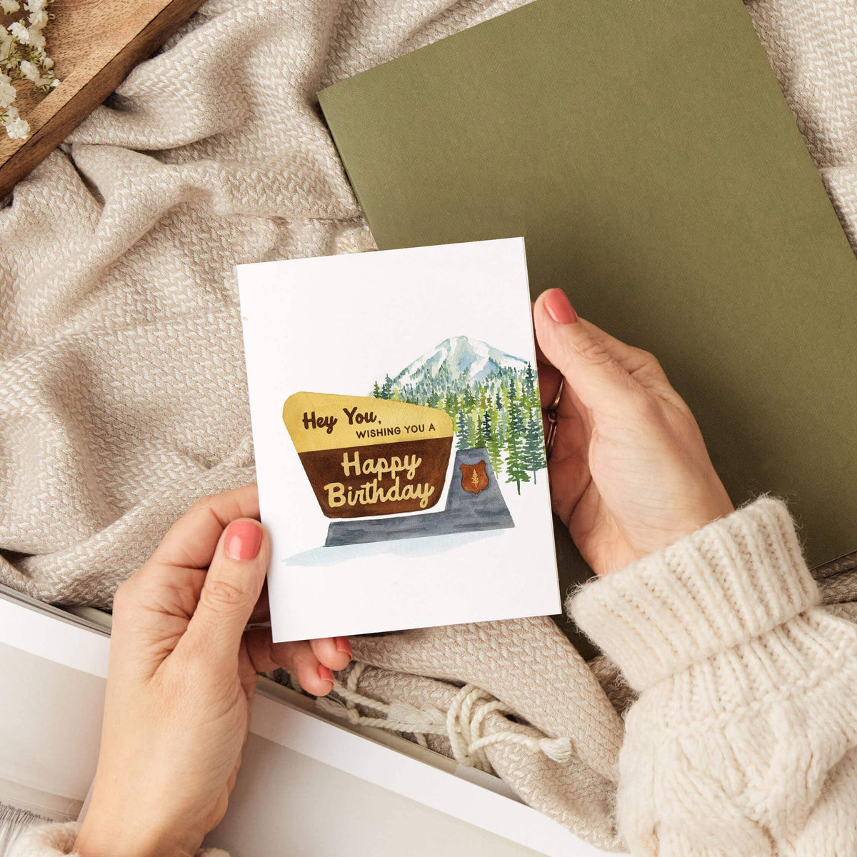 Hands holding the Hey You National Park Birthday Card over a cozy scene with a knit sweater, woven blanket, and an olive green notebook