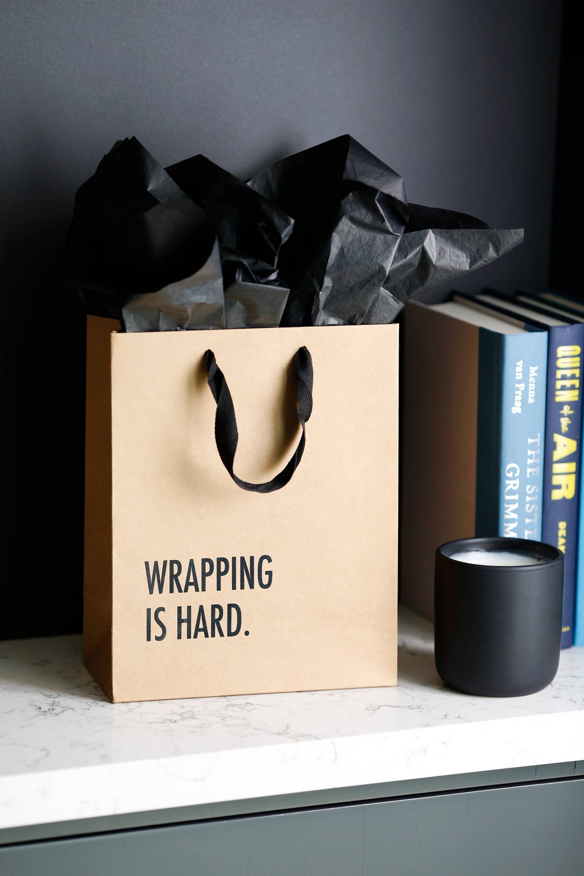 Kraft gift bag printed WRAPPING IS HARD. with black tissue paper, on a marble shelf beside books and a black candle