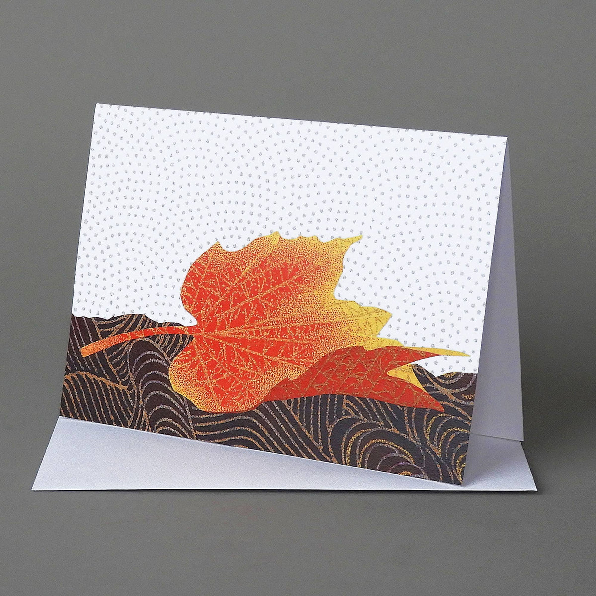 Fallen Maple notecard of an orange and yellow maple leaf resting on dark swirled origami paper, on an envelope