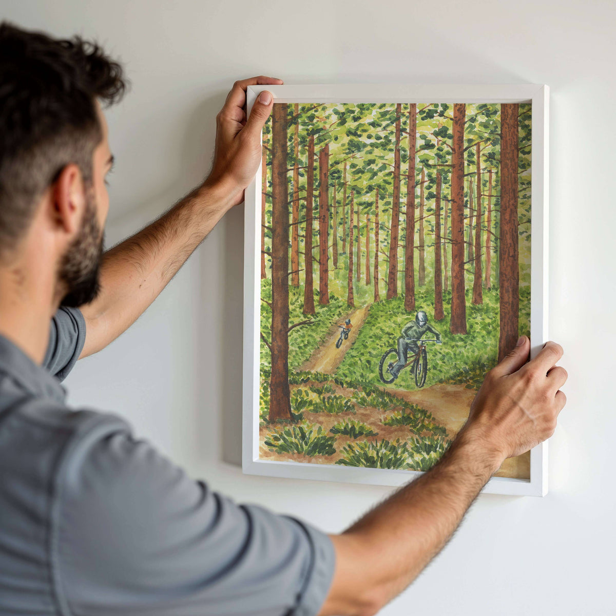 Person hanging the framed Pedaling Through the Pines Watercolor Art Print in a white frame on a white wall