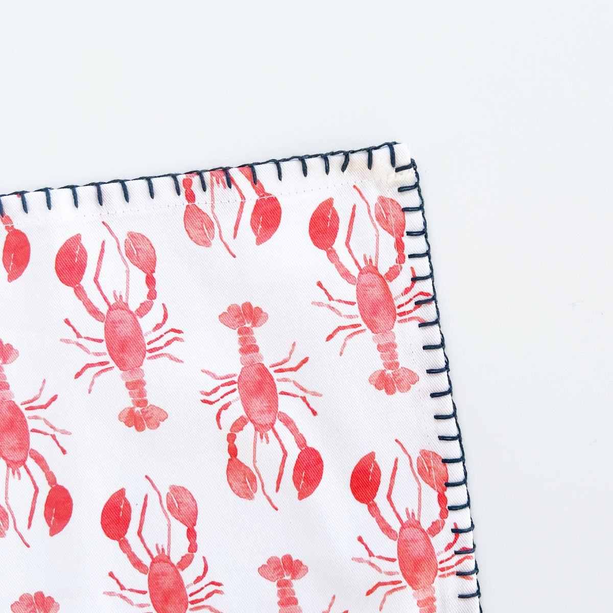 Corner of a lobster napkin showing red watercolor lobsters and navy whipstitch embroidery