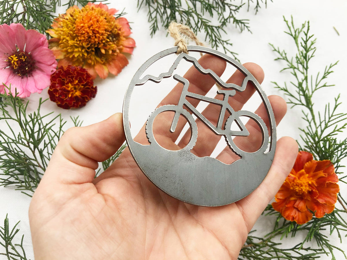 Raw steel mountain bike ornament held in an open hand to show its size, with orange zinnias and juniper behind