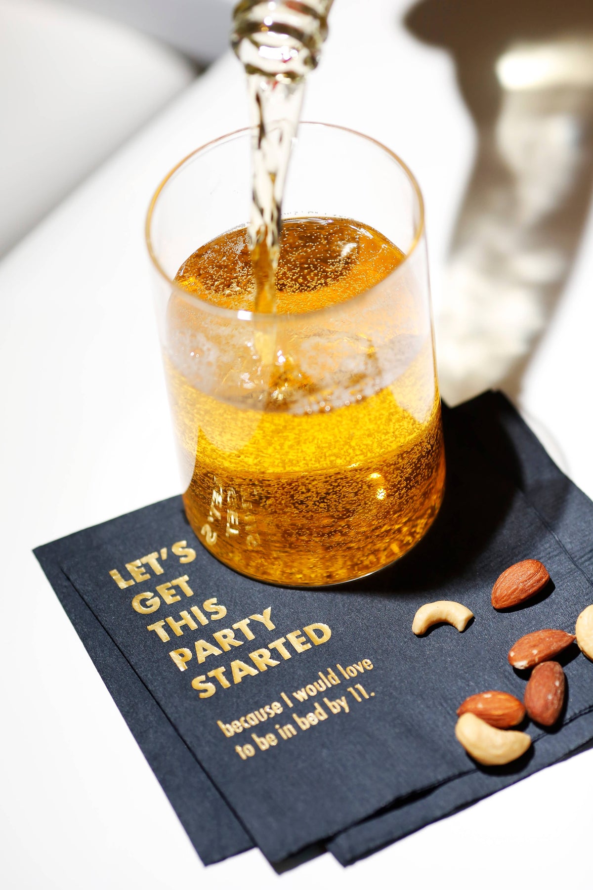 Black cocktail napkins with gold foil text under a glass of sparkling drink being poured, beside nuts, on white