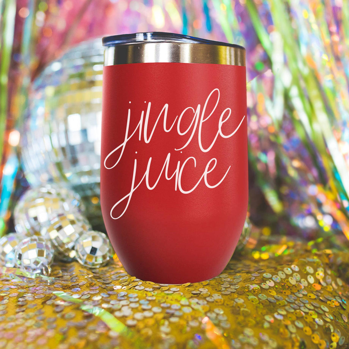 Red Jingle Juice stainless steel tumbler with white script lettering on a gold sequin cloth beside mirror disco balls