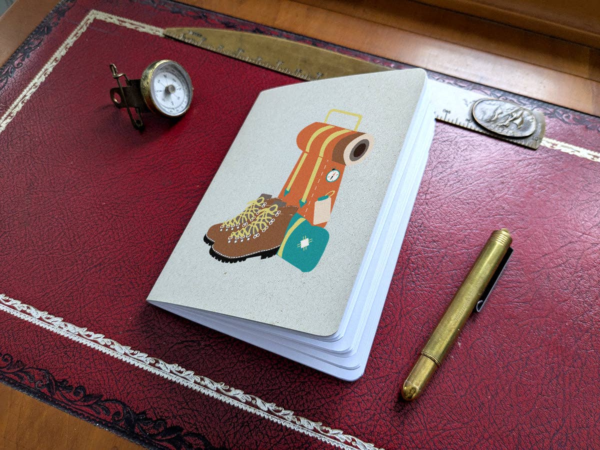 Thru-Hiker pocket notebook with a kraft cover printed with an orange backpack and brown hiking boots, on a red leather desk pad with a compass and brass pen