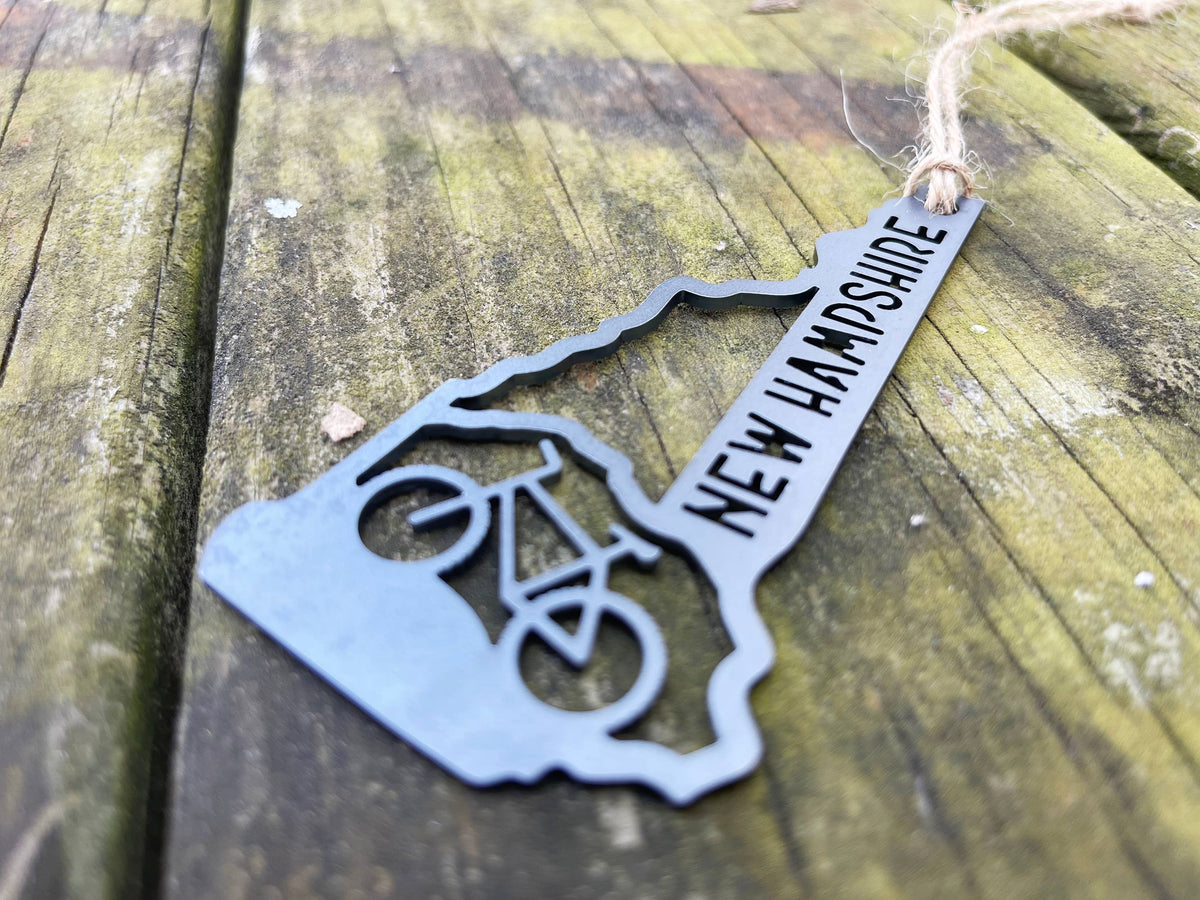 Raw steel New Hampshire mountain bike ornament seen at a low angle, with a cut-out bike and twine loop, on mossy wood