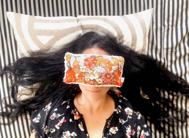 Woman with long black hair lying on striped bedding wearing an orange and cream retro floral eye pillow over her eyes