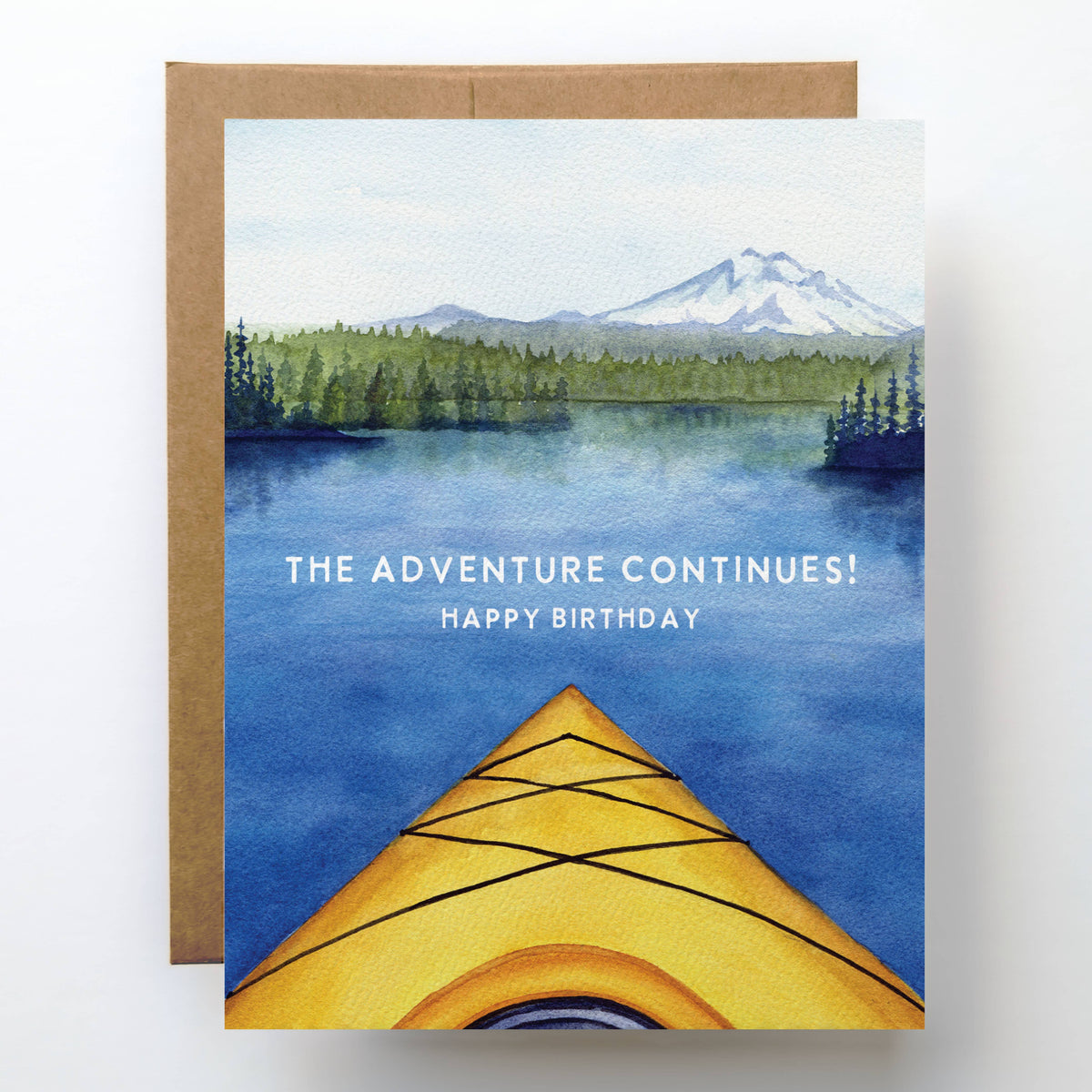 Kayak Birthday Card showing the view from a yellow kayak bow across a calm alpine lake toward a snow capped mountain, text reads The Adventure Continues Happy Birthday, kraft envelope behind
