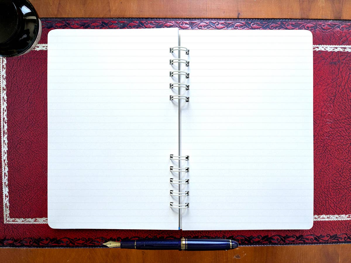 Autumn Biking spiral notebook open flat to lined pages, with a navy fountain pen and an inkwell on a red leather desk pad
