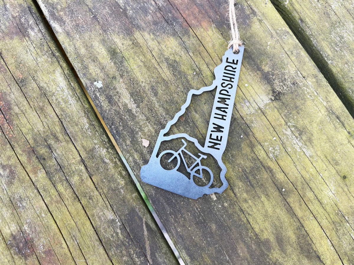 Raw steel New Hampshire mountain bike ornament with a bike cutout and twine hanger, lying diagonally on mossy wood boards