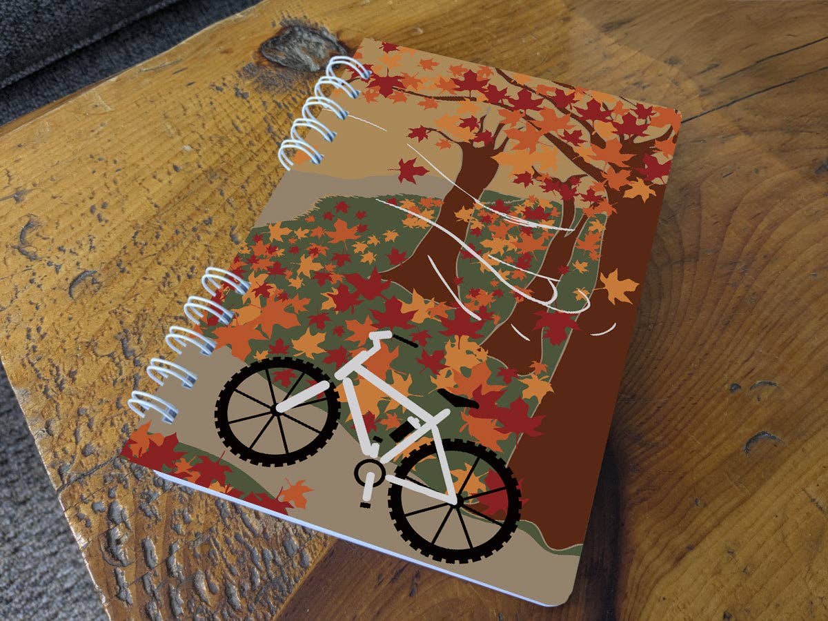 Autumn Biking spiral notebook resting on a wooden table, its cover showing a white mountain bike on a trail among autumn maple trees