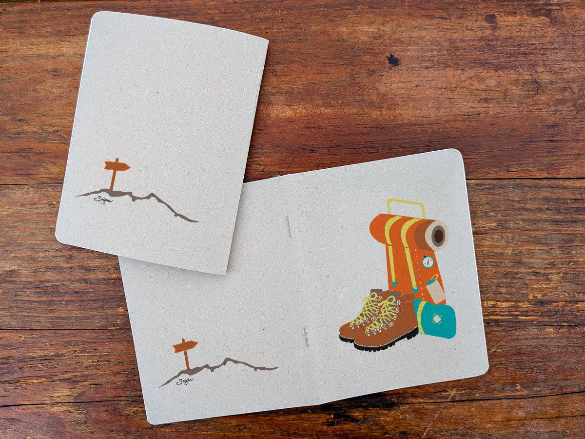 Staple-bound Thru-Hiker notebook with a trail marker sign on a ridge on the back cover, open beside the front cover with backpack and boots