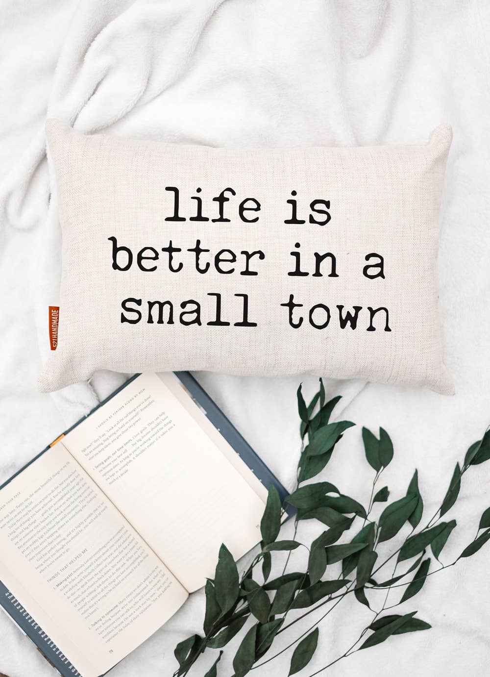 Life is Better in a Small Town Linen Pillow lying flat on a white blanket next to an open book and dried eucalyptus sprigs