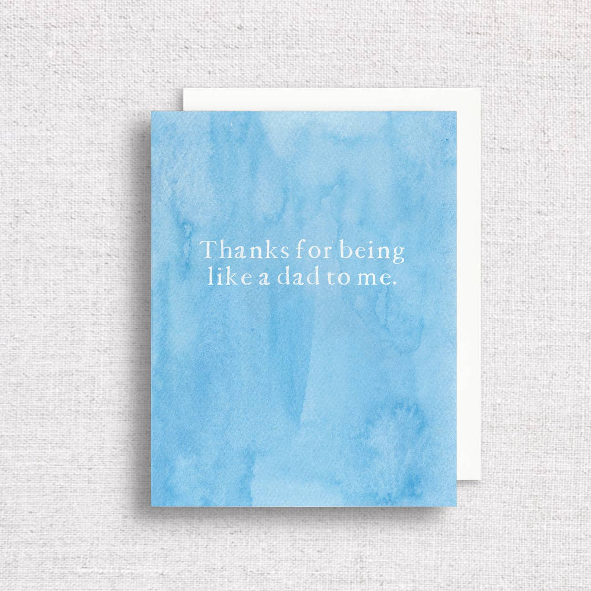 Thanks for being like a dad to me in white serif type on a watercolor blue card, on a white envelope over linen