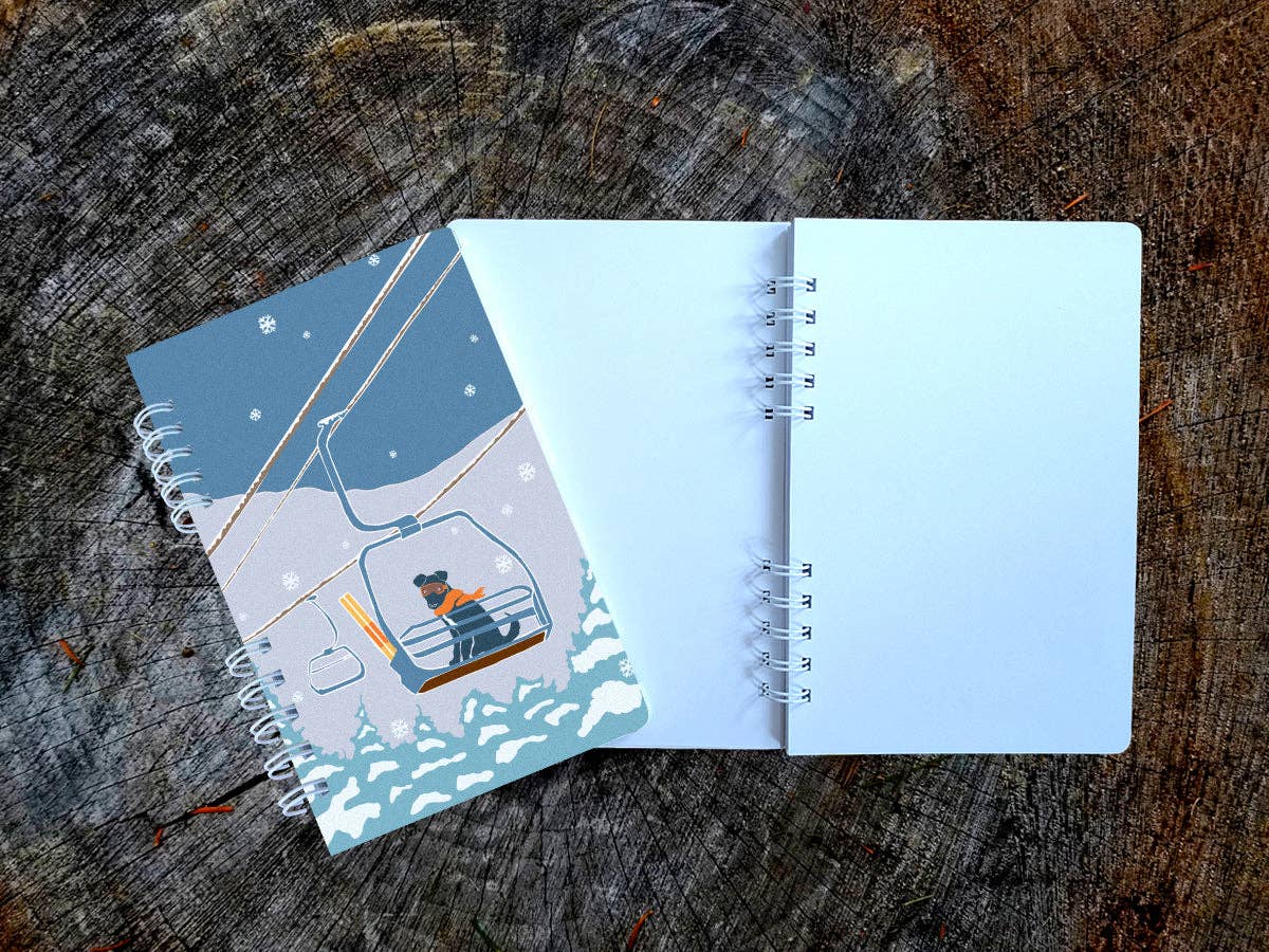 Ski Lift Pup spiral notebook with a pup in an orange scarf riding a snowy chairlift on the cover, open to a blank page, on a tree stump