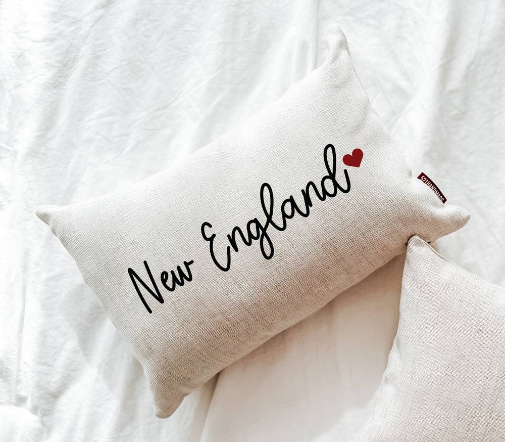 Cream linen lumbar pillow with New England in black script and a small red heart, on white bedding beside a second pillow