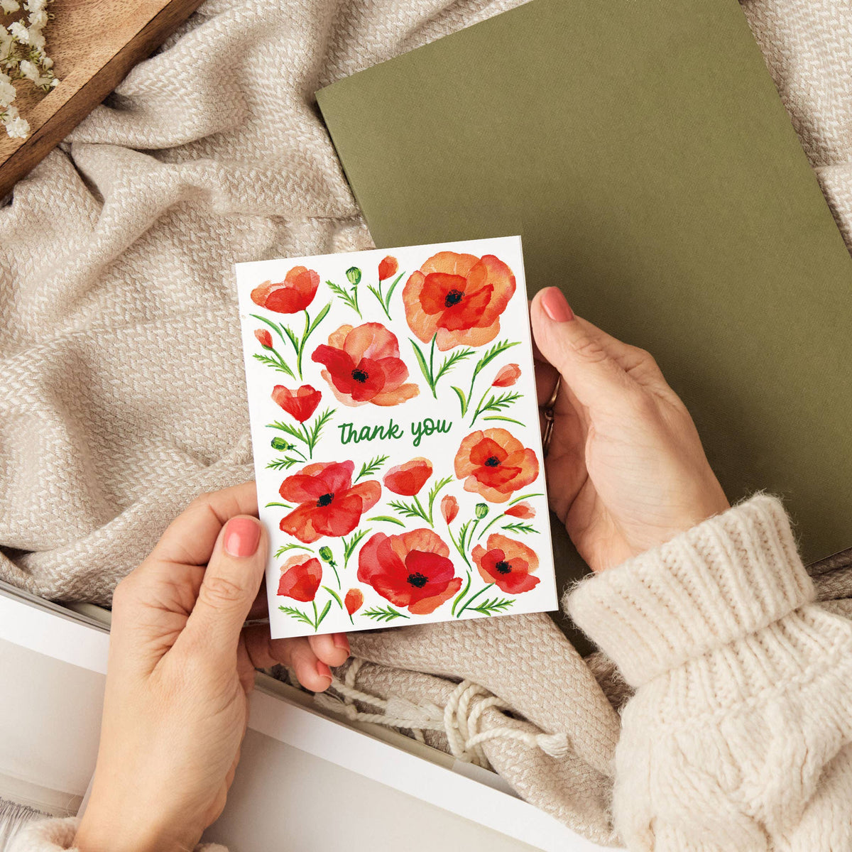 Hands holding the Poppy Thank You Card over a cozy scene with a knit blanket and an olive green notebook
