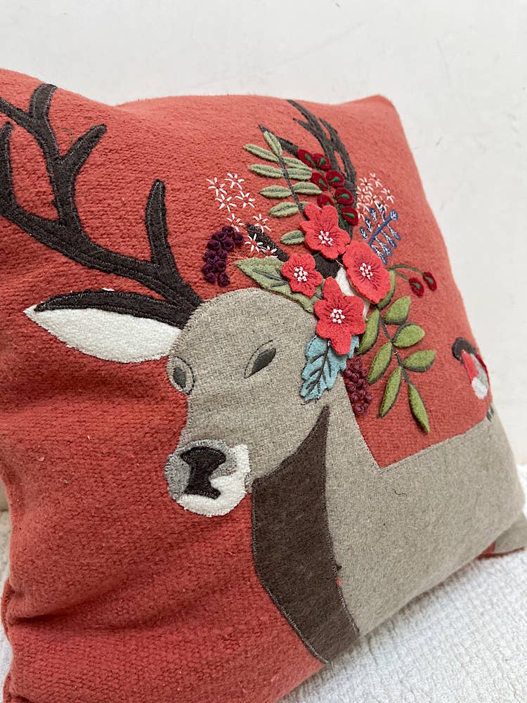 Angled close-up of a rust red pillow with an appliqued gray felt deer, red felt flowers, leaves and berries