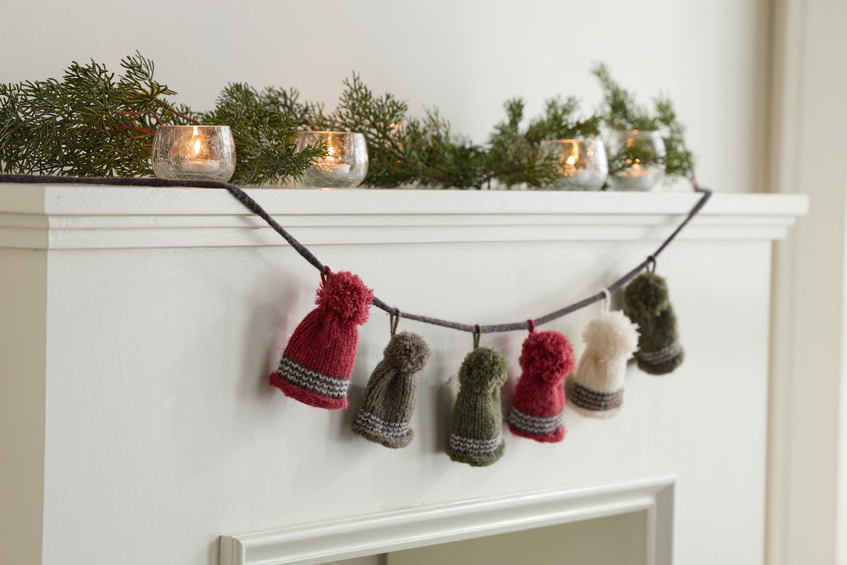 Garland of tiny pom-pom knit hats in red, green, gray and cream hung above a white mantel with candles