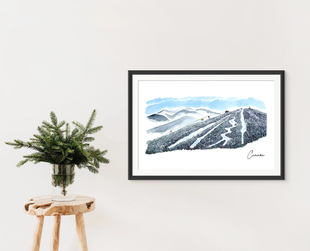 Cannon Mountain trail view print in a black frame on a wall, styled with a wooden stool and a vase of pine branches