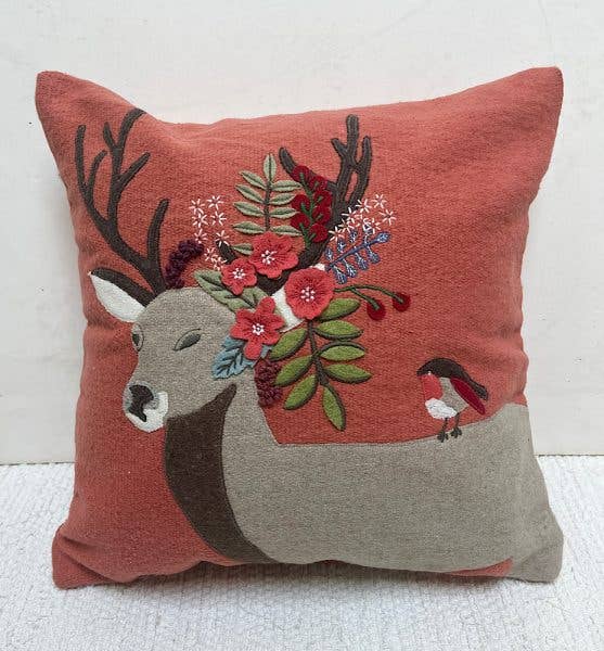 Rust red square pillow with a felt-appliqued deer with a bouquet of red flowers and leaves on its antlers and a robin