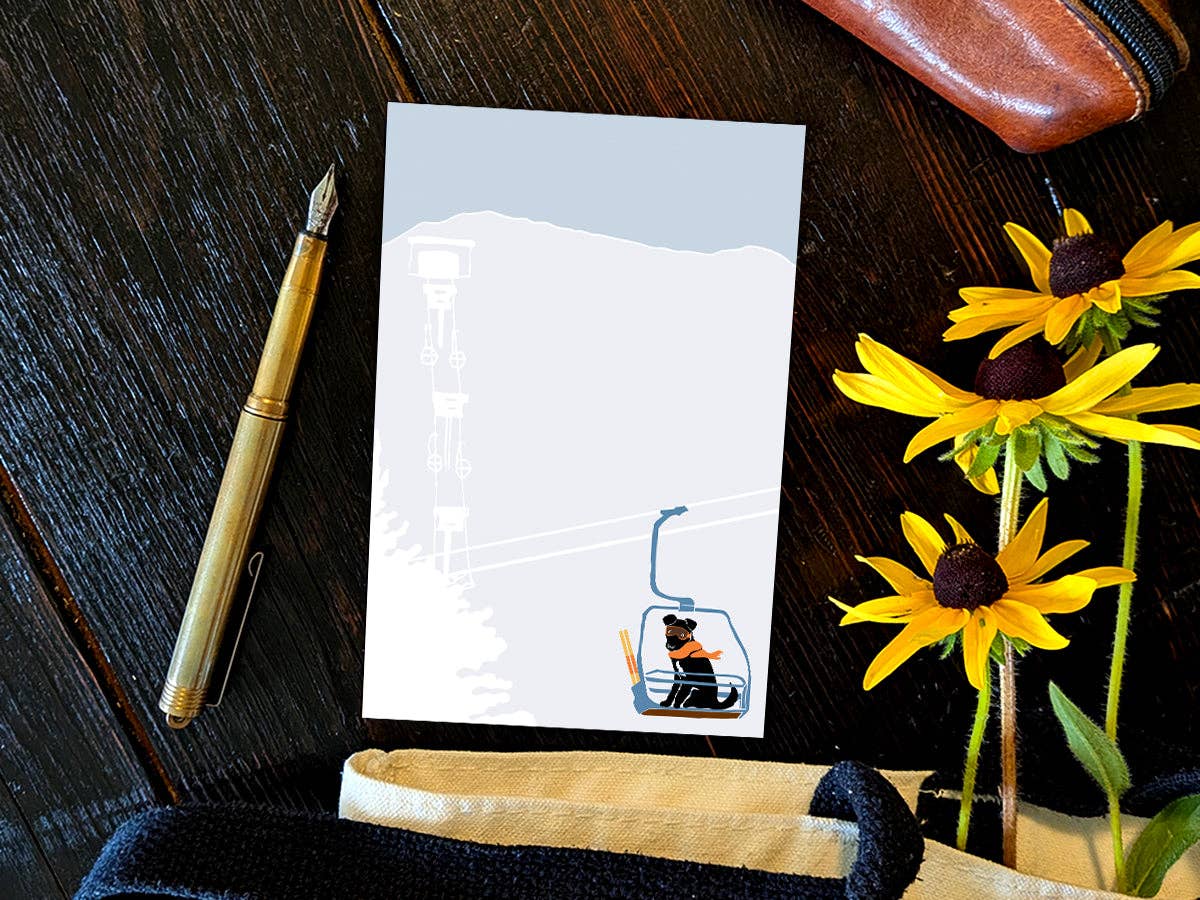 Ski Lift Pup notepad on a dark wood desk beside a brass fountain pen, a leather pencil case, and yellow black-eyed Susan flowers