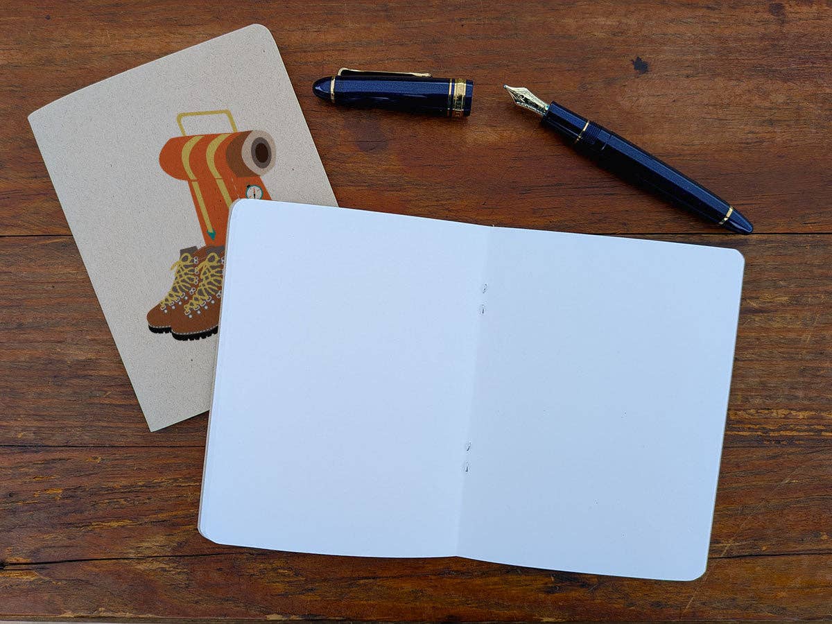 Thru-Hiker pocket notebook open to blank white pages beside its kraft cover, with a navy fountain pen and its cap on a wood table