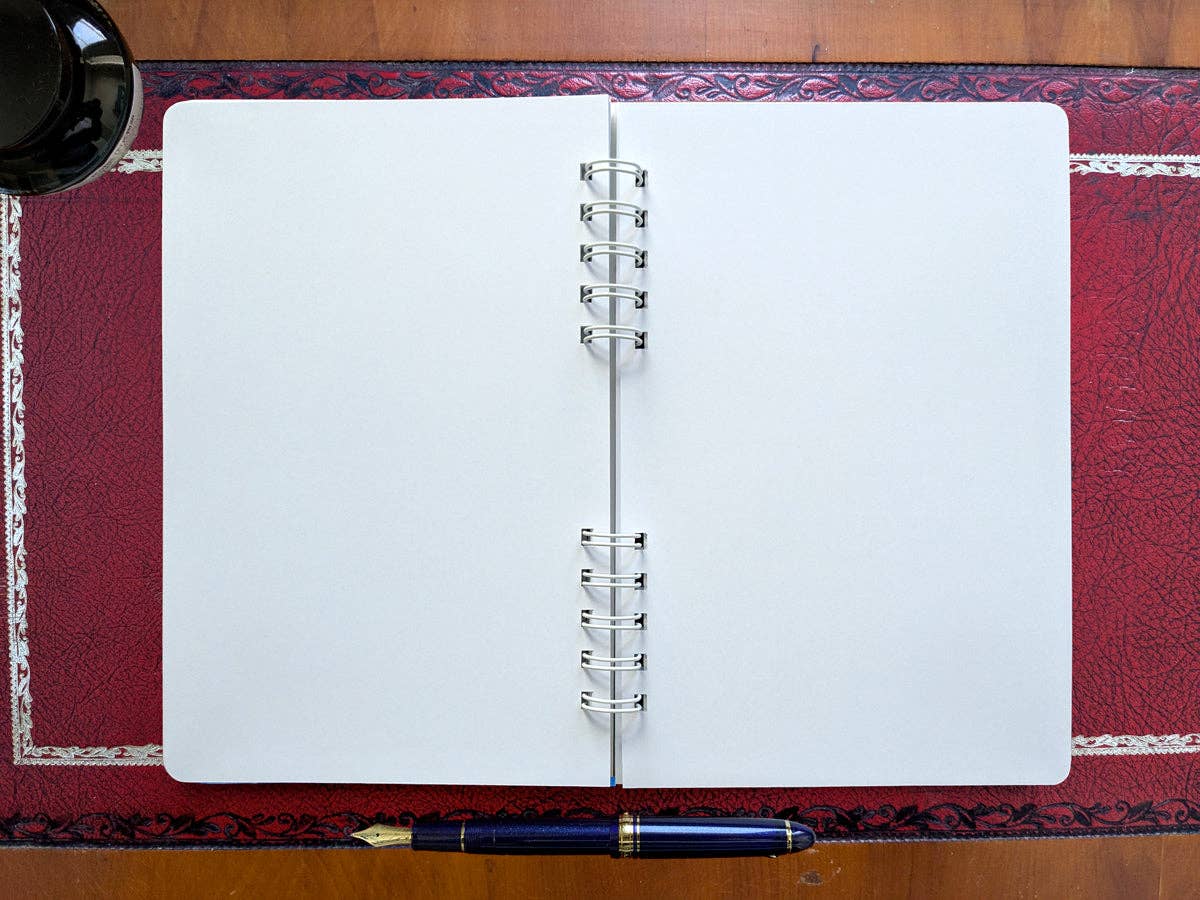 Autumn Biking spiral notebook open flat to blank, unlined pages, with a navy fountain pen and an inkwell on a red leather desk pad