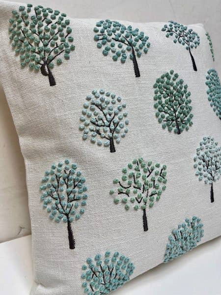 Angled close-up of a white cotton pillow embroidered with round-crowned trees in blue-green and gray french knots