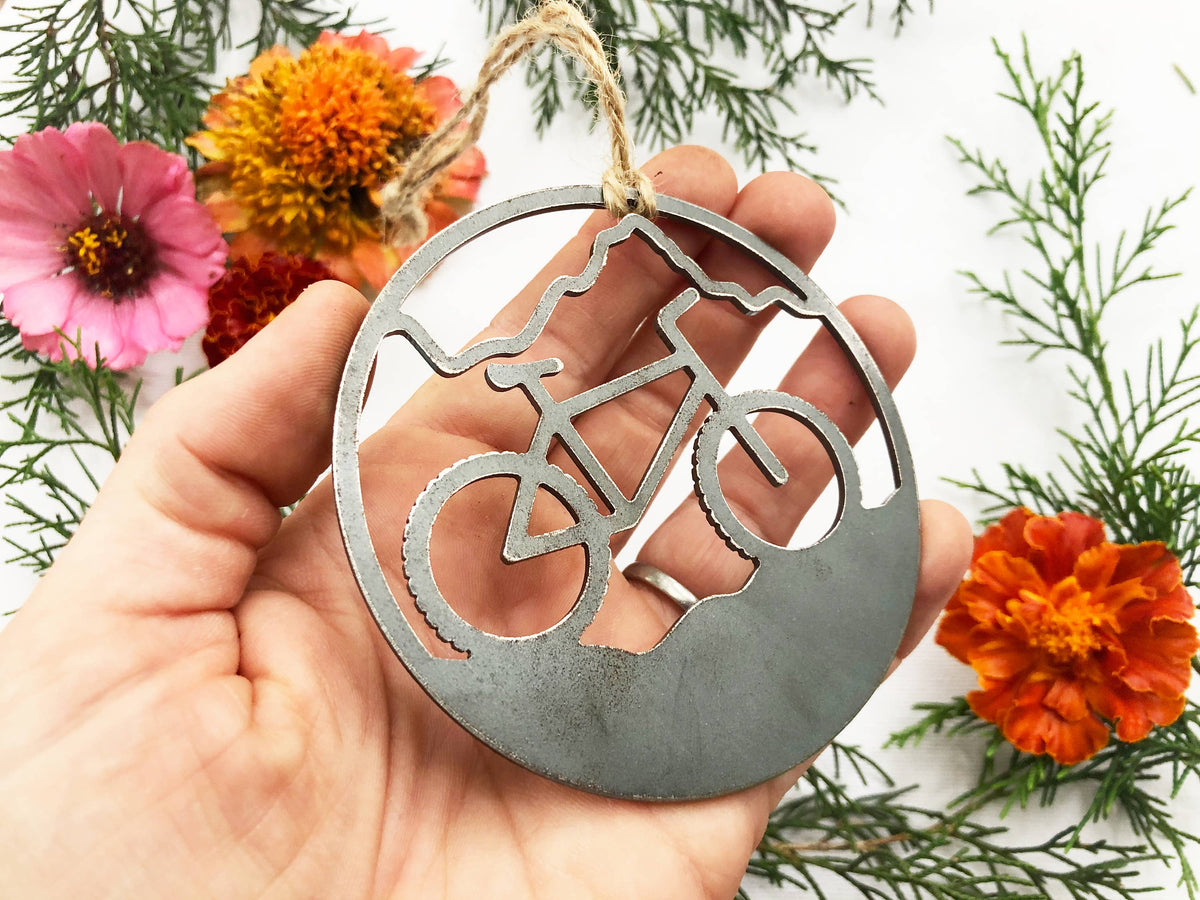 Hand holding a raw steel mountain bike ornament, a round cutout with a bike and mountain ridge, among flowers