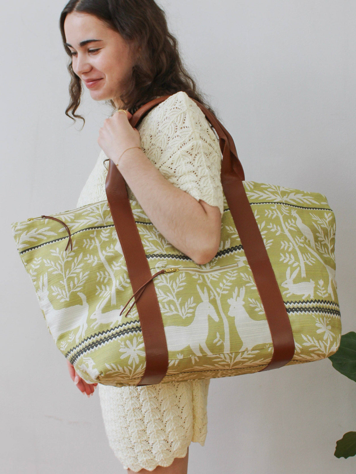 A woman carrying a Folk Life Carry On Travel Bag over her shoulder, green and cream jacquard fabric with rabbit, deer, and bird motifs, brown leather straps