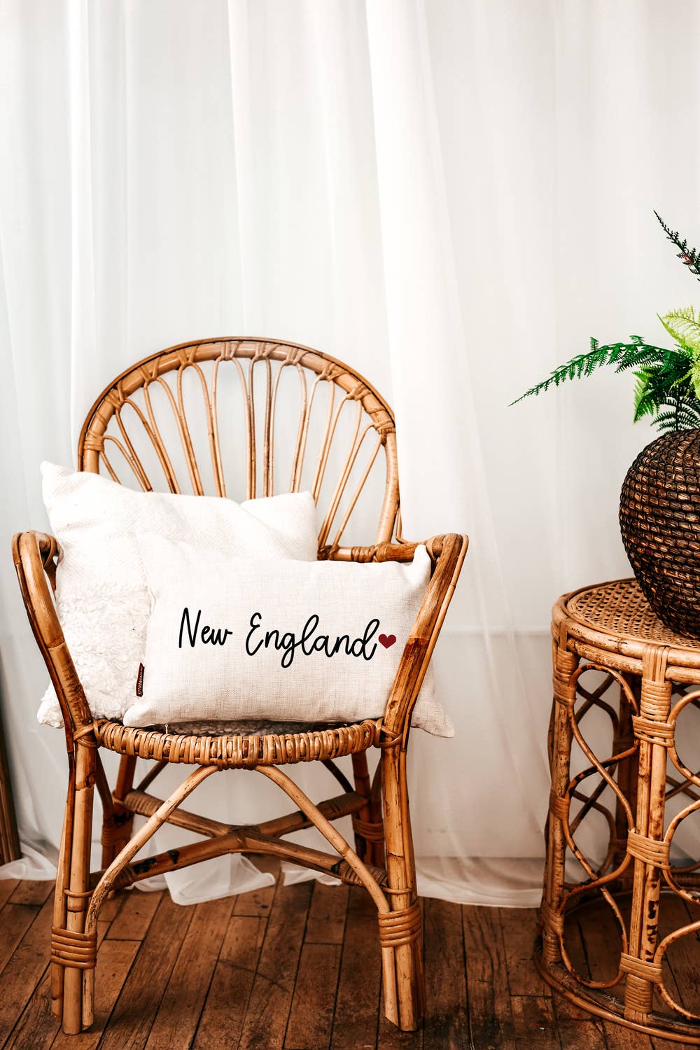 New England linen lumbar pillow with black script and a red heart on a wicker chair in front of sheer white curtains