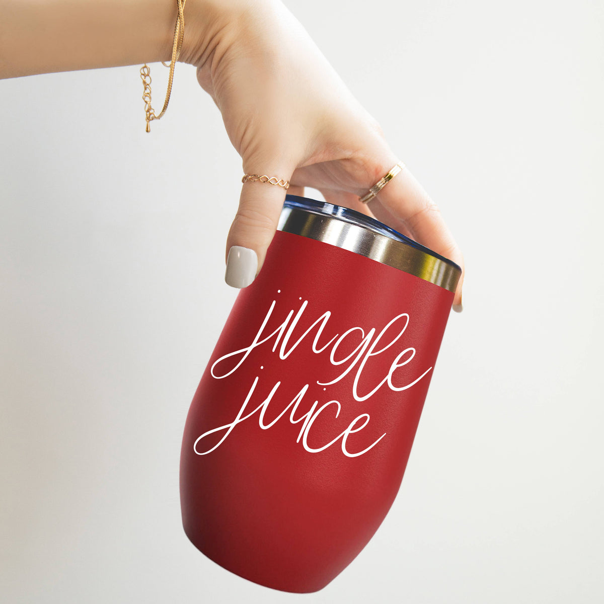 Hand tilting a red stainless steel tumbler with jingle juice lettered in white script, light gray backdrop