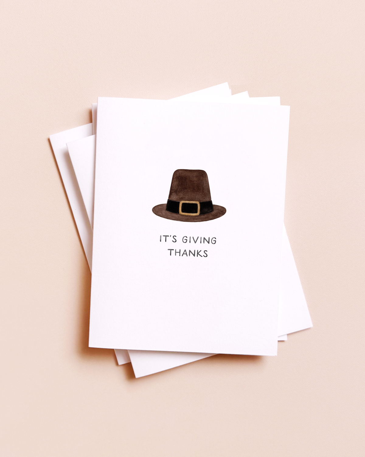 Stack of white greeting cards with a watercolor pilgrim hat and the words IT'S GIVING THANKS on a peach background