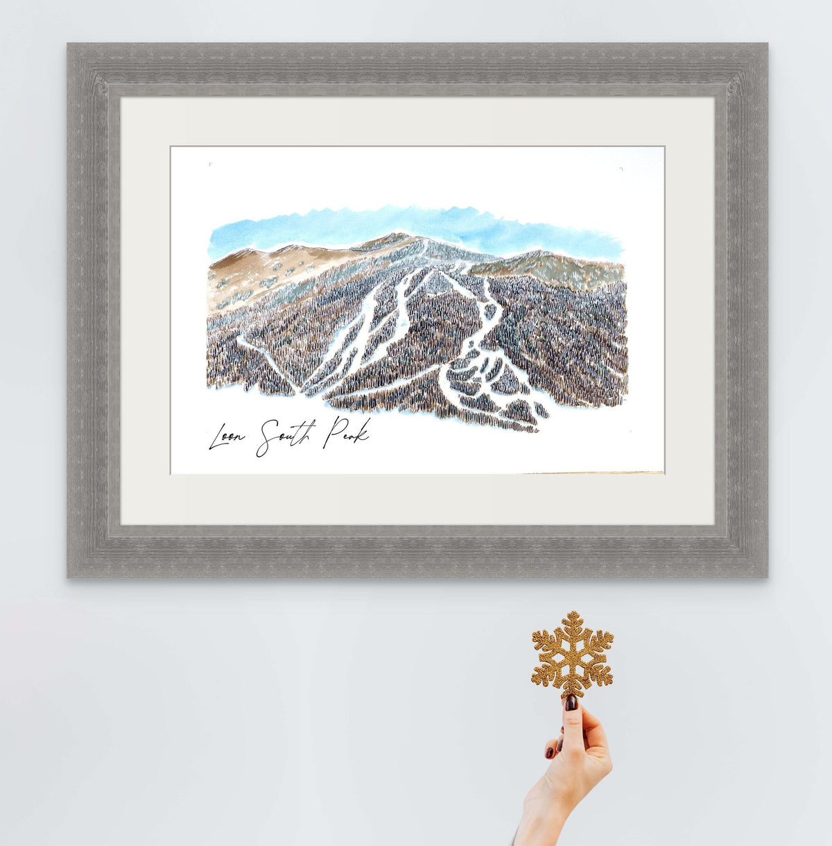 Loon South Peak print in a grey textured frame, styled with a hand holding a glitter snowflake ornament