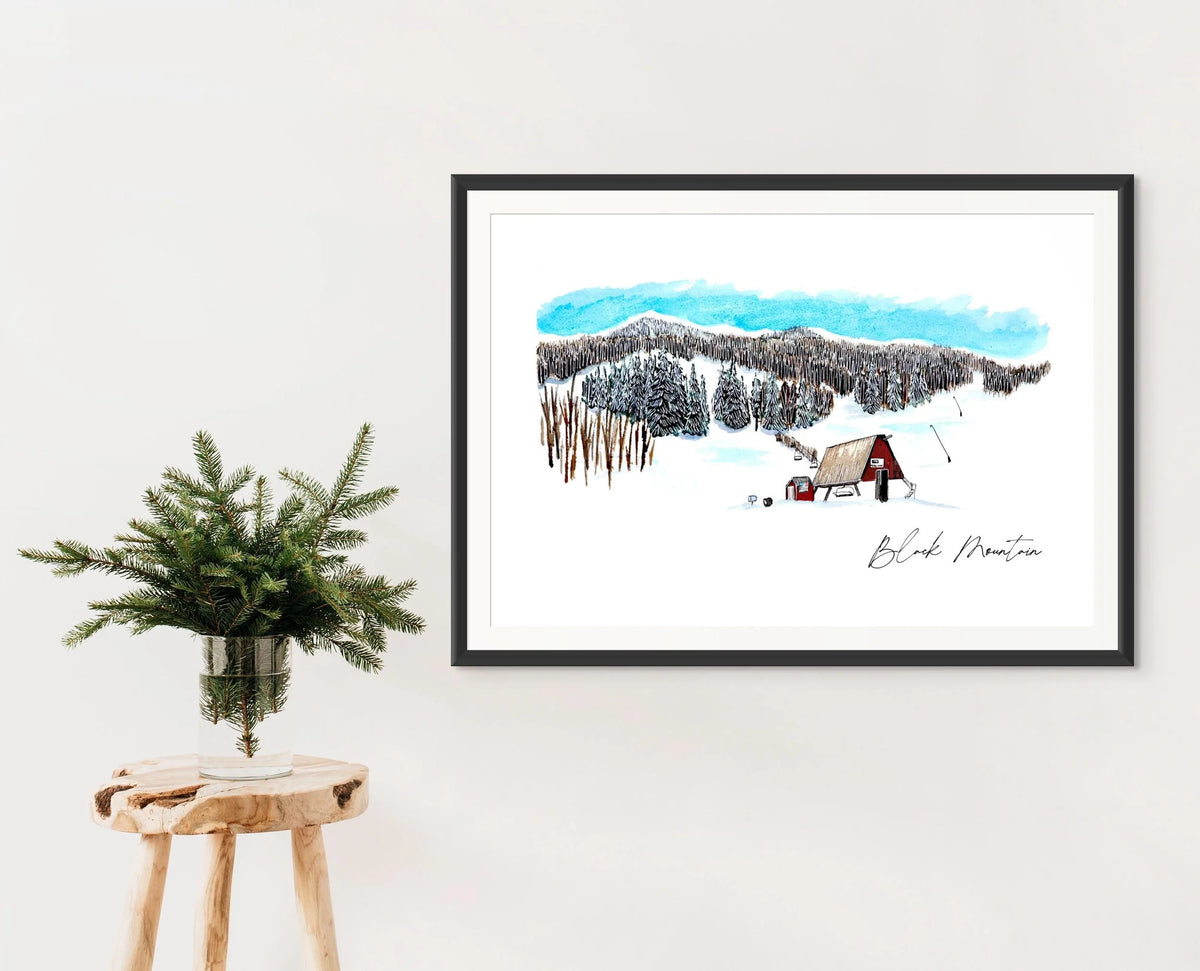 Black Mountain Ski Area print in a black frame on a wall, styled with a wooden stool and a vase of pine branches
