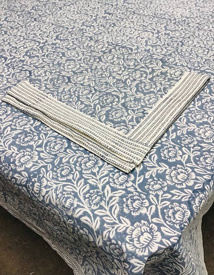 Block-printed napkin with a dotted stripe border folded across a matching blue-gray peony tablecloth