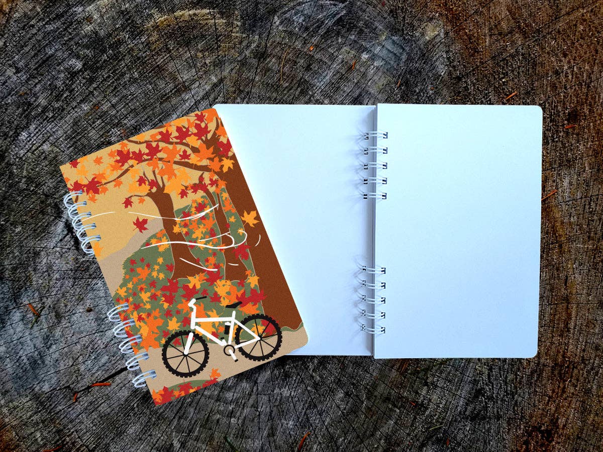 Autumn Biking spiral notebook with a mountain bike on a leaf-covered forest trail under red and orange maples, open to a blank page, on a tree stump