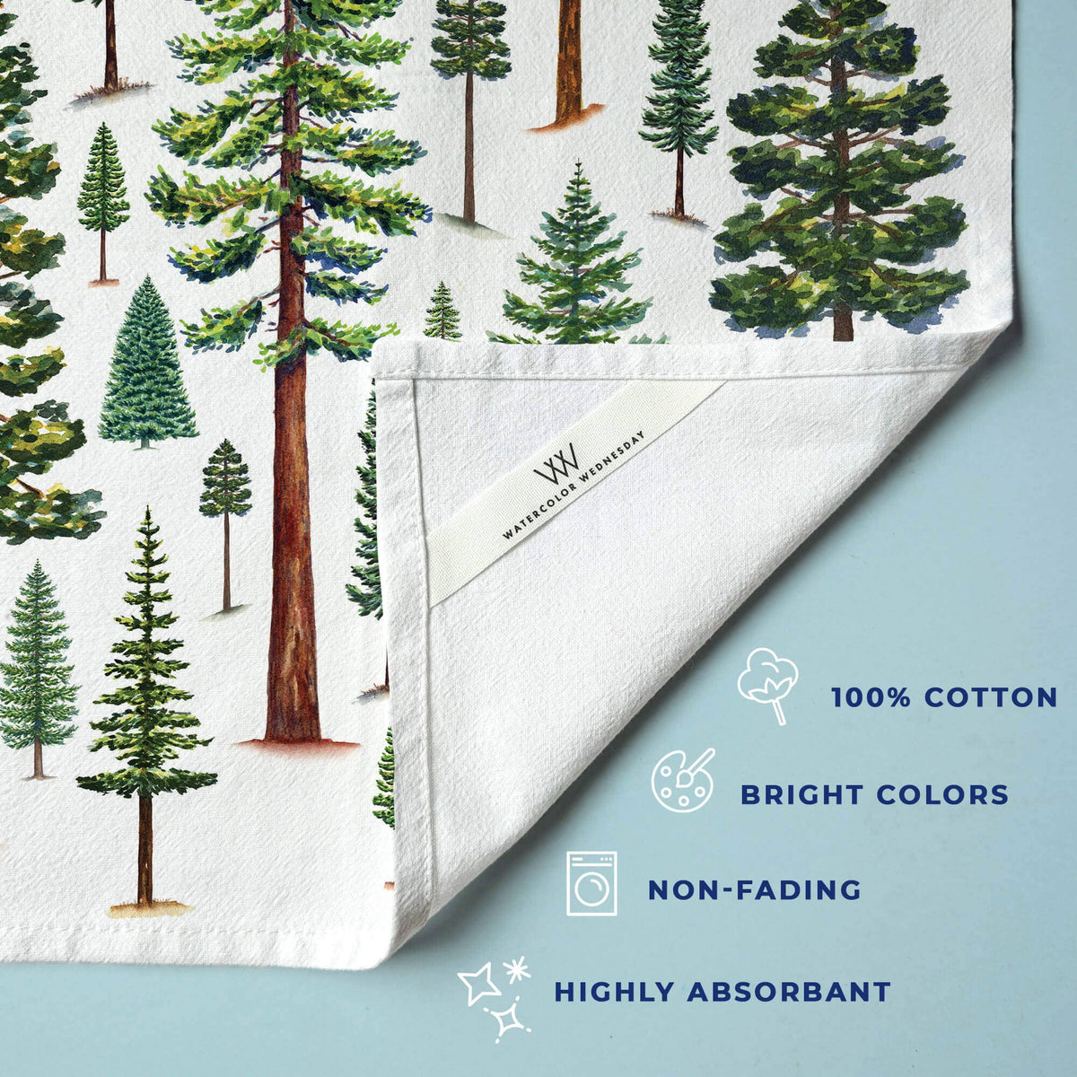 Close-up of Evergreen Trees Tea Towel corner showing pine and fir trees, with the Watercolor Wednesday label and 100% cotton, bright colors, non-fading, highly absorbant feature icons